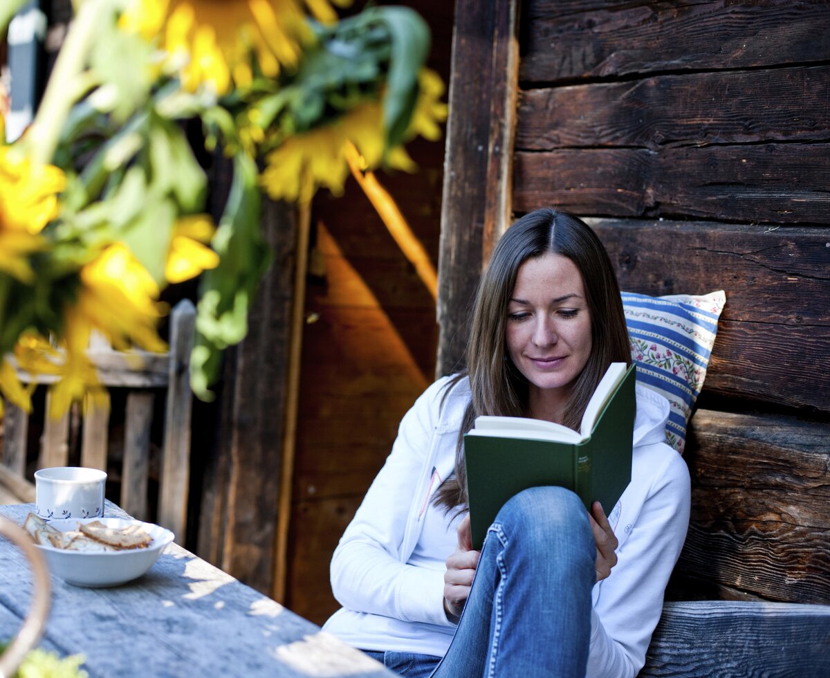 Frau sitzt mit einem Buch auf der Bank vor der Almhütte, Steiermark | © Urlaub am Bauernhof Steiermark / Kufferath